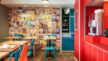 A cafe interior with colorful chairs, tables, a wall of travel posters, and a red counter with a coffee machine.