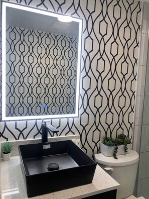 Un baño con un lavamanos negro, un espejo con luces LED, un inodoro y un revestimiento de pared estampado.