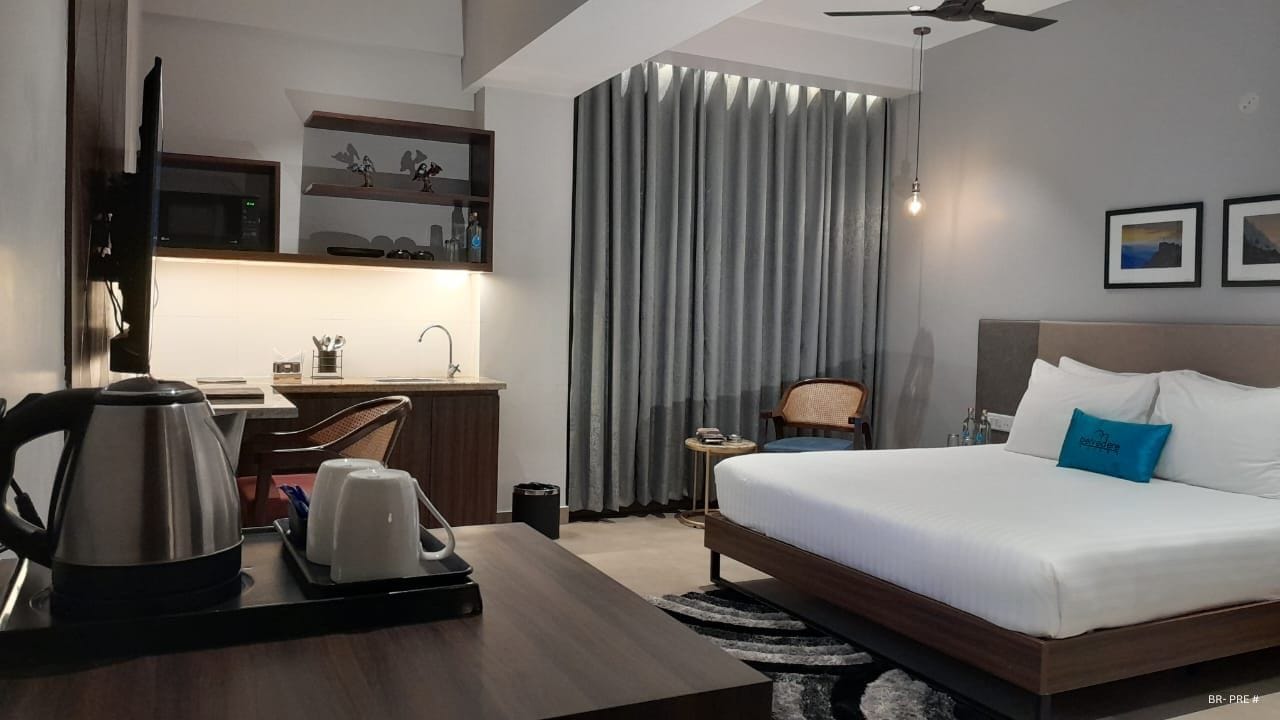 A modern hotel room with a bed, desk, and kitchenette.