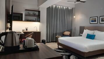 A modern hotel room with a bed, desk, and kitchenette.
