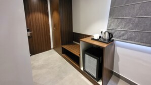 Kompakti hotellihuone, jossa on pieni keittiönurkkaus, pieni jääkaappi ja oleskelualue, jossa on penkki.
