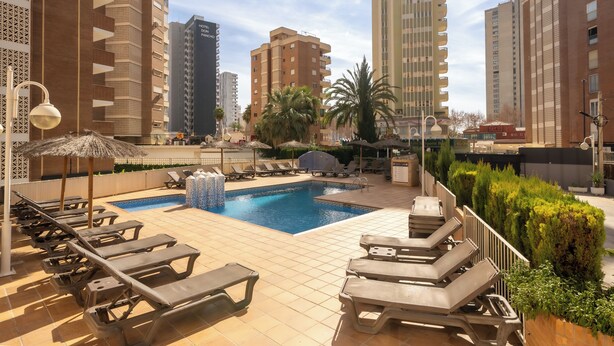 Área de piscina no terraço com espreguiçadeiras, piscina e edifícios altos ao fundo.