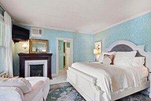 A bedroom with a white bed, a fireplace, a television, and a door leading to another room.