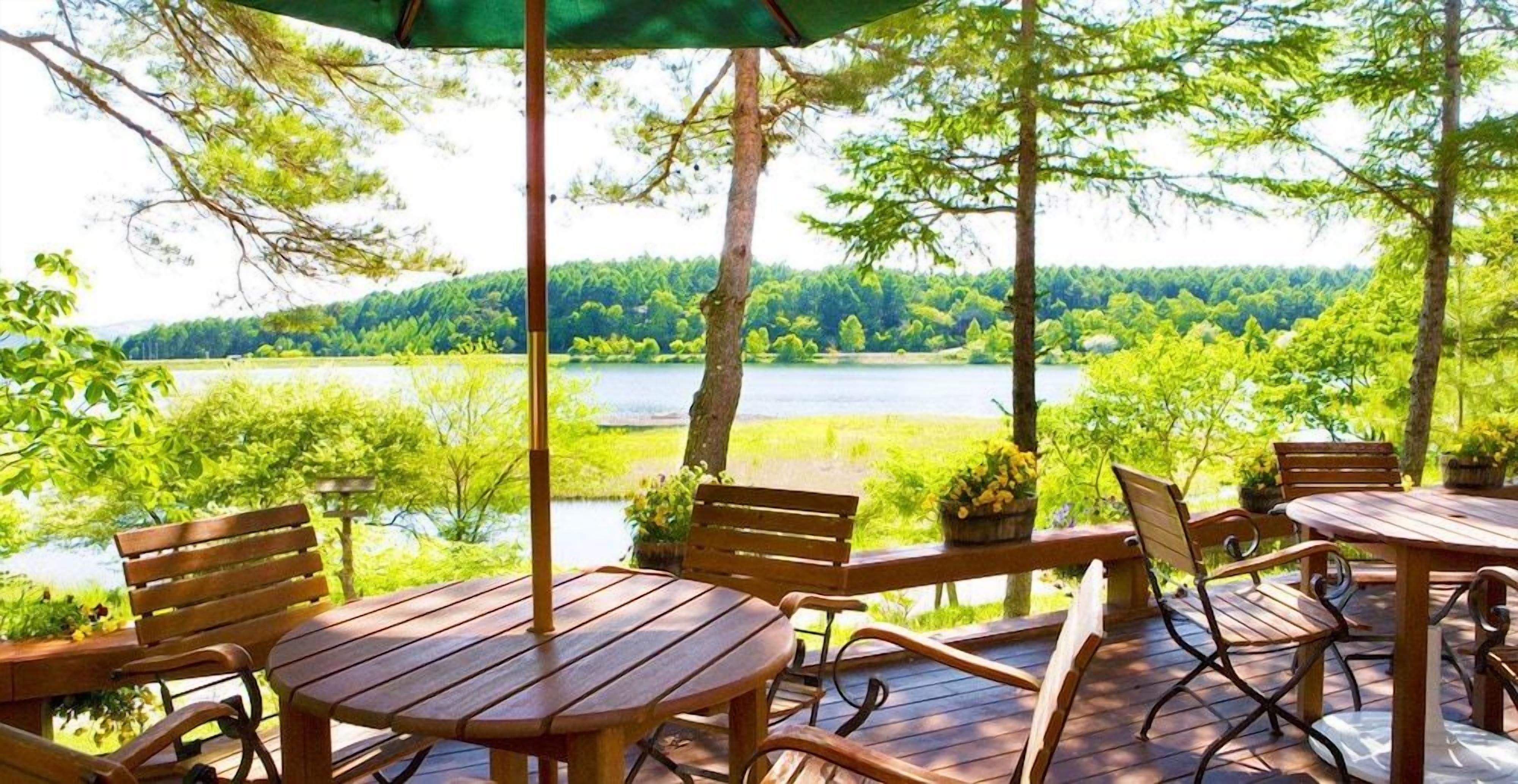 An outdoor patio with wooden tables and chairs, surrounded by trees and overlooking a lake.