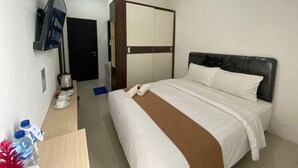 A hotel room with a bed, a TV mounted on the wall, a desk with a kettle, and a wardrobe. - Amaze Hotel Kediri (Ngasem)