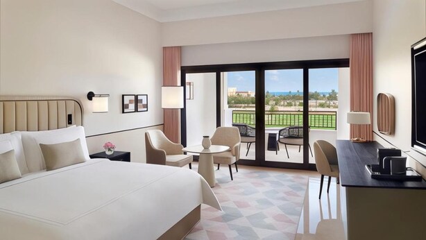 Quarto de hotel com uma cama grande, uma escrivaninha, uma cadeira, uma mesa pequena e uma varanda com vista.