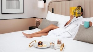 Uma pessoa deitada na cama de um quarto de hotel, lendo um livro. O quarto possui uma obra de arte emoldurada na parede, uma mesa de cabeceira com abajur e um serviço de mesa com comida e bebidas sobre a cama.