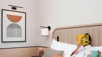 Een persoon ligt in een hotelkamer op bed, een boek lezend. De kamer heeft een lijst met kunst aan de muur, een nachtkastje met een lamp, en een dienblad met eten en drinken op het bed.