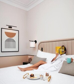 Une personne est allongée sur un lit dans une chambre d’hôtel et lit un livre. La chambre est décorée avec un tableau encadré au mur, une table de chevet avec une lampe, et une table d’appoint sur le lit contenant de la nourriture et des boissons.