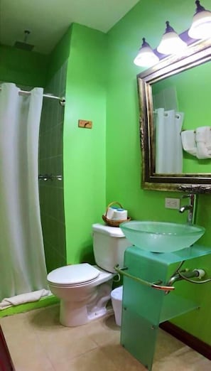 A bathroom with a green wall, a white toilet, a glass sink, a shower curtain, and a mirror with lights. - Hotel Royal Blue (San Pedro)