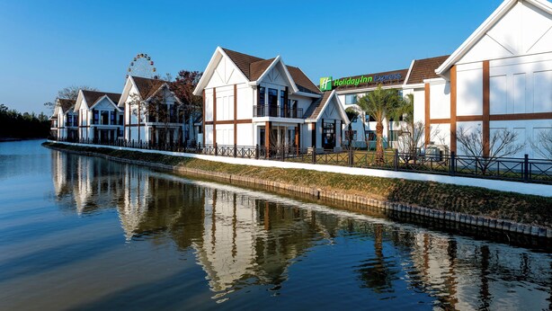 Ein Hotel am Wasser mit einem Riesenrad im Hintergrund, einem Holiday Inn Express Schild und einem ruhigen Gewässer, das die Gebäude spiegelt.