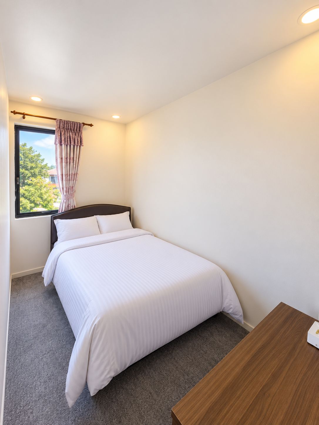 Una cama individual con cabecera de madera, una ventana con cortina y una pequeña mesa de madera.