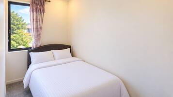 Una cama individual con cabecera de madera, una ventana con cortina y una pequeña mesa de madera.