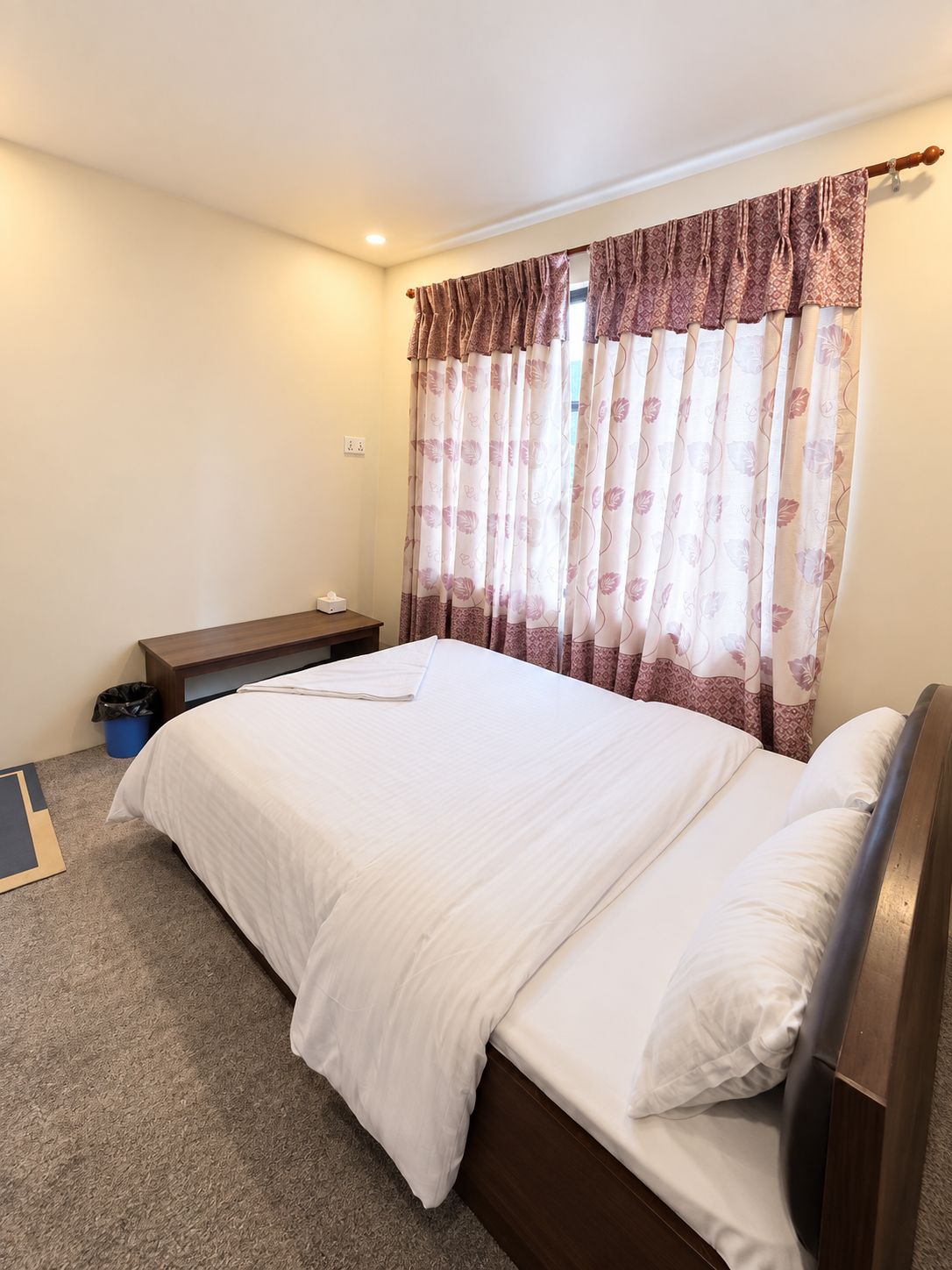 A bedroom with a single bed, a bedside table, a trash bin, and a window with curtains.