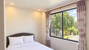 Una cama bien hecha con sábanas blancas, una ventana con cortinas y vistas al verdor exterior.