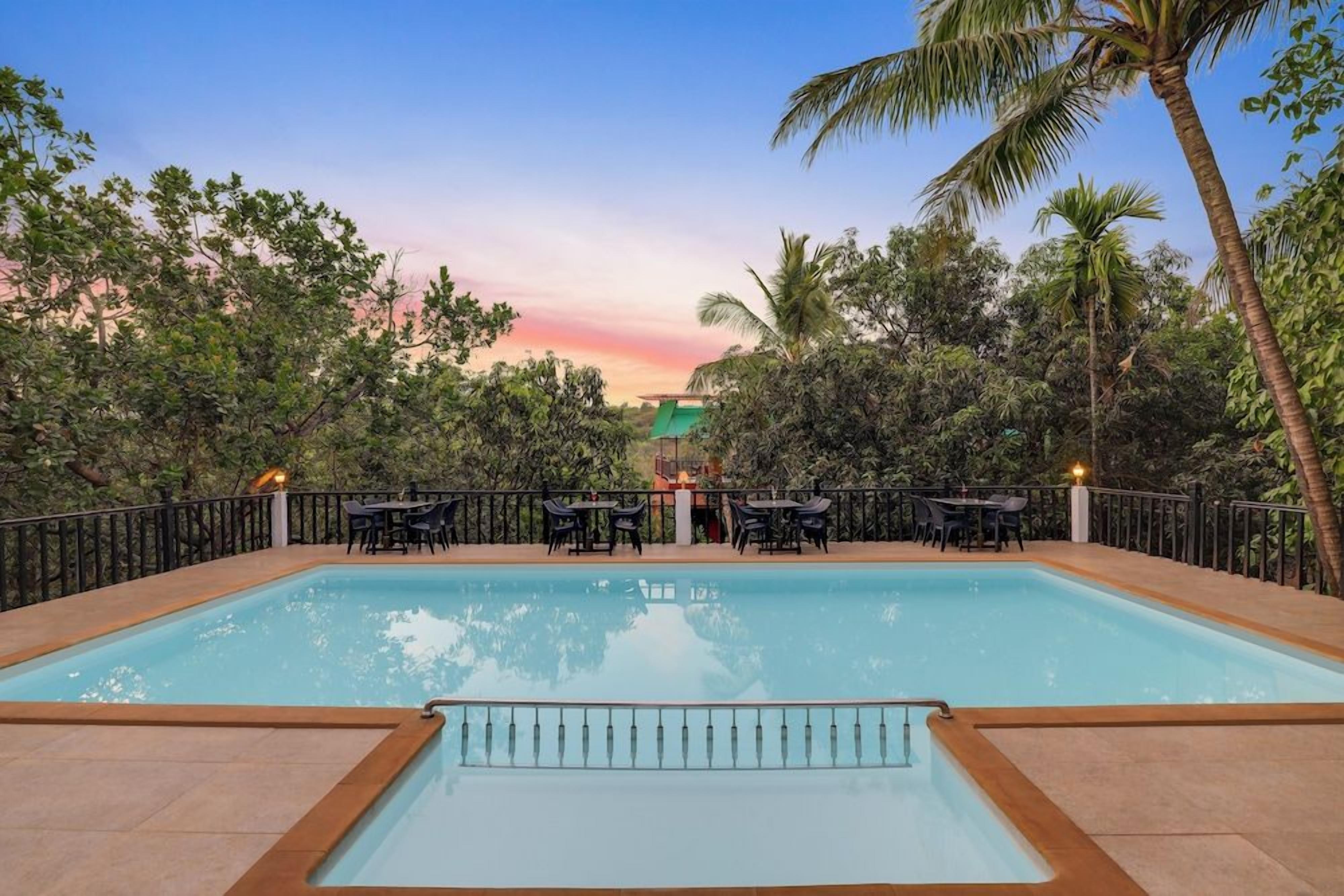 Una piscina rectangular con un deck alrededor, rodeada de árboles y un atardecer al fondo.