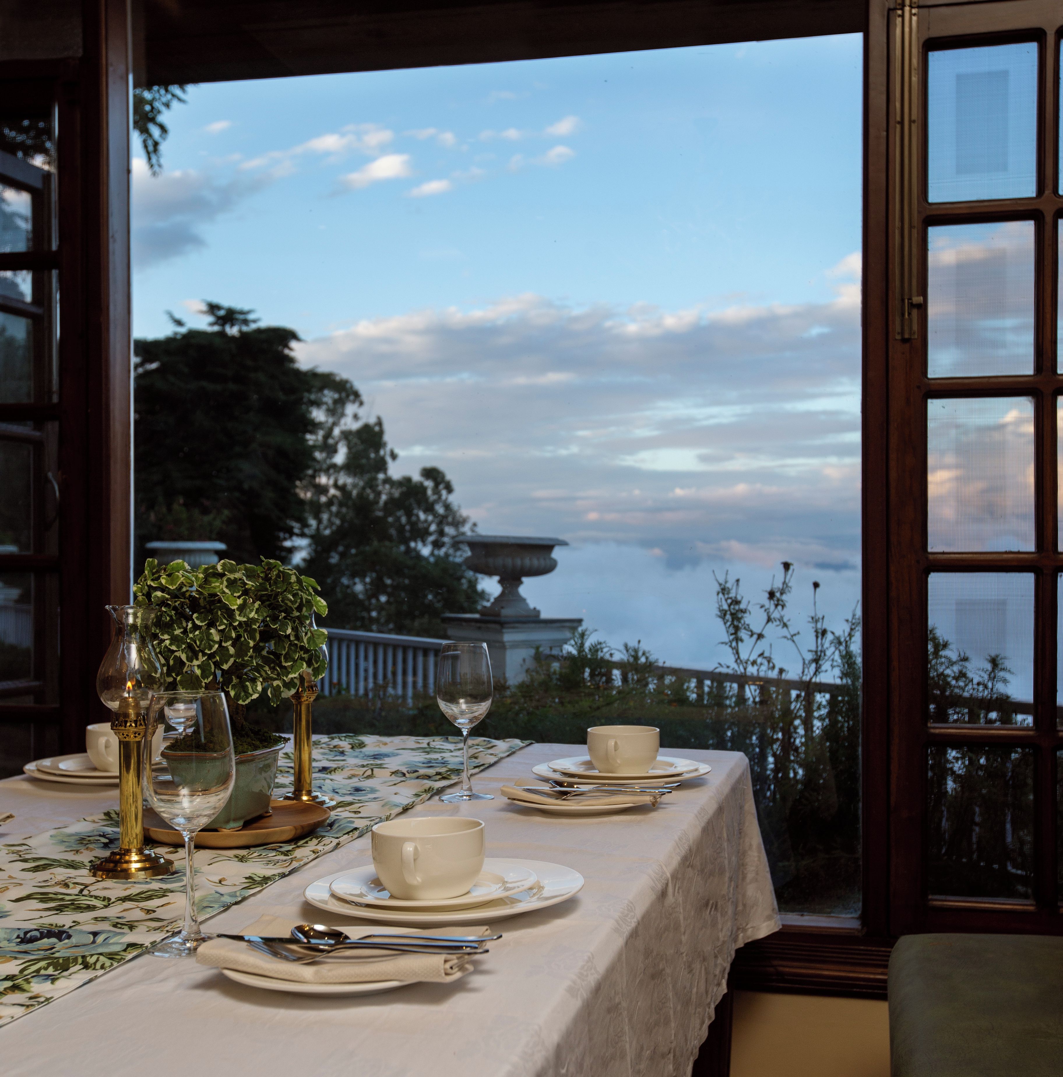 Una mesa de comedor con platos, tazas y cubiertos, con vistas a un paisaje con árboles y un edificio.