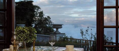 A dining table set with plates, cups, and cutlery, overlooking a scenic view with trees and a building.