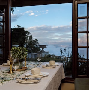 A dining table set with plates, cups, and cutlery, overlooking a scenic view with trees and a building. - Daleside Manor (Kasauli)