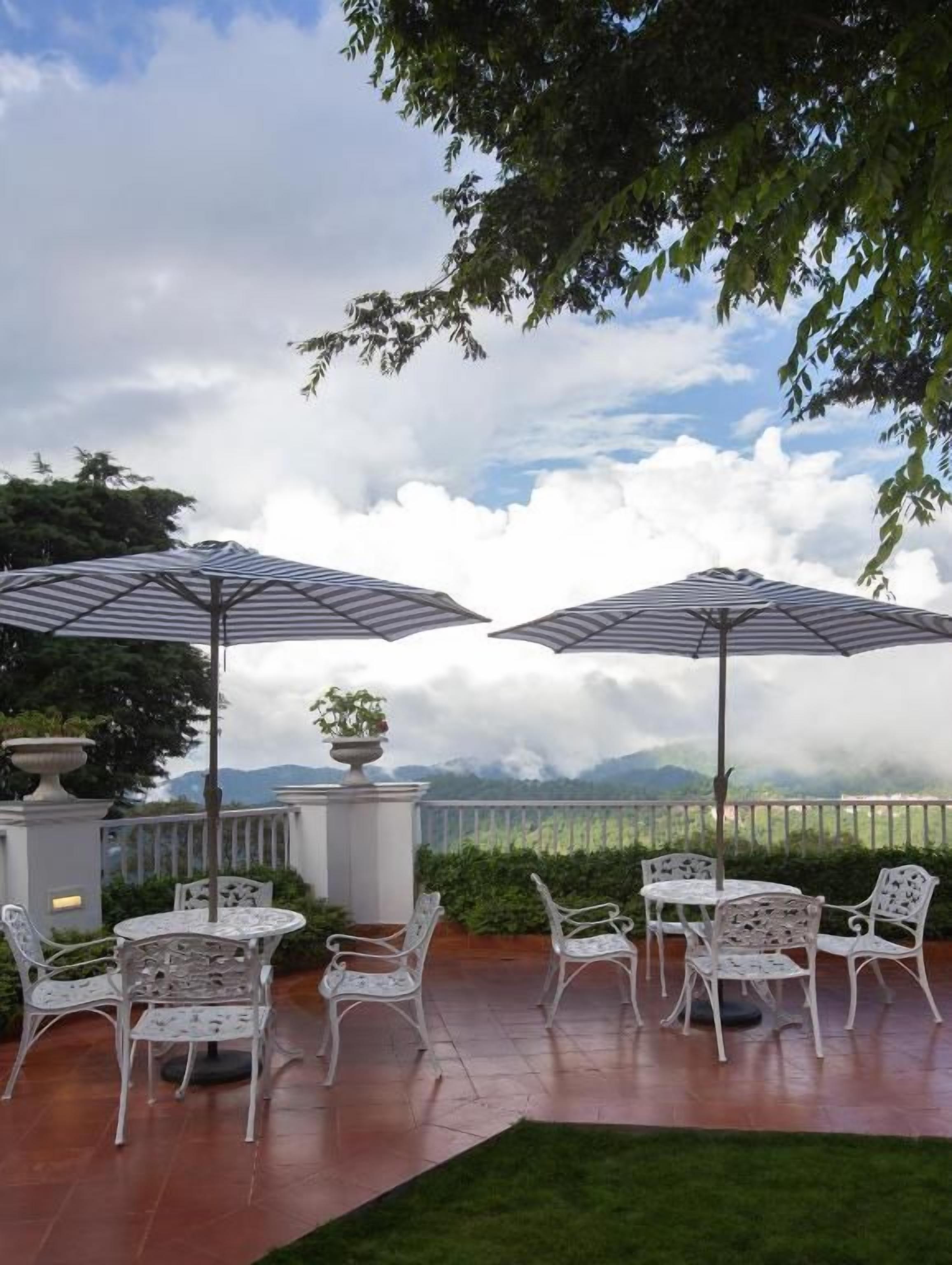 Un patio exterior con sillas y mesas de metal blanco, dos sombrillas y una vista de montañas al fondo.