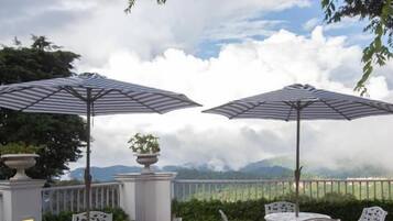 Un patio exterior con sillas y mesas de metal blanco, dos sombrillas y una vista de montañas al fondo.