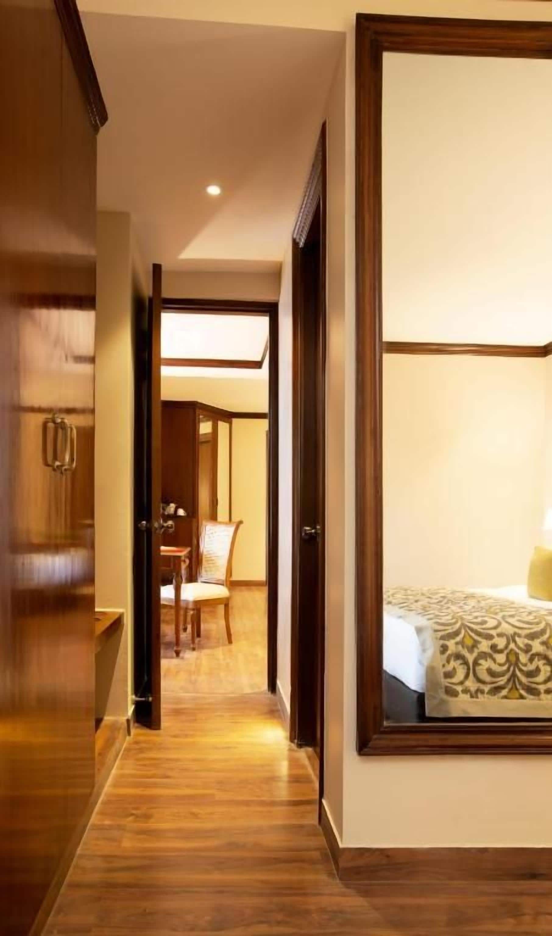 A hotel hallway with a wooden floor, a bed with patterned bedding, and a chair in the background.