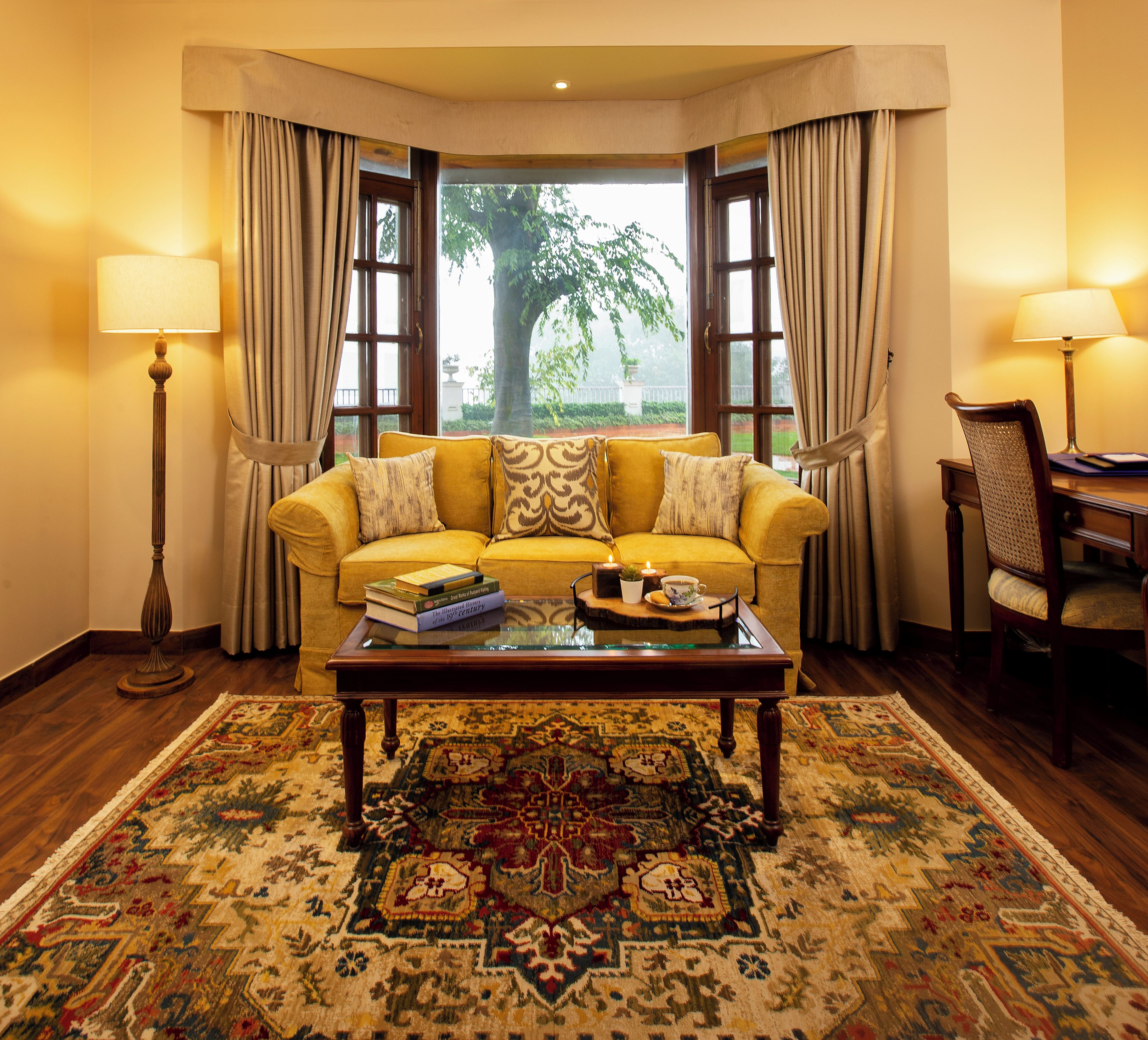 A living room with a yellow sofa, a glass coffee table, a patterned rug, and a view of a tree through a window.
