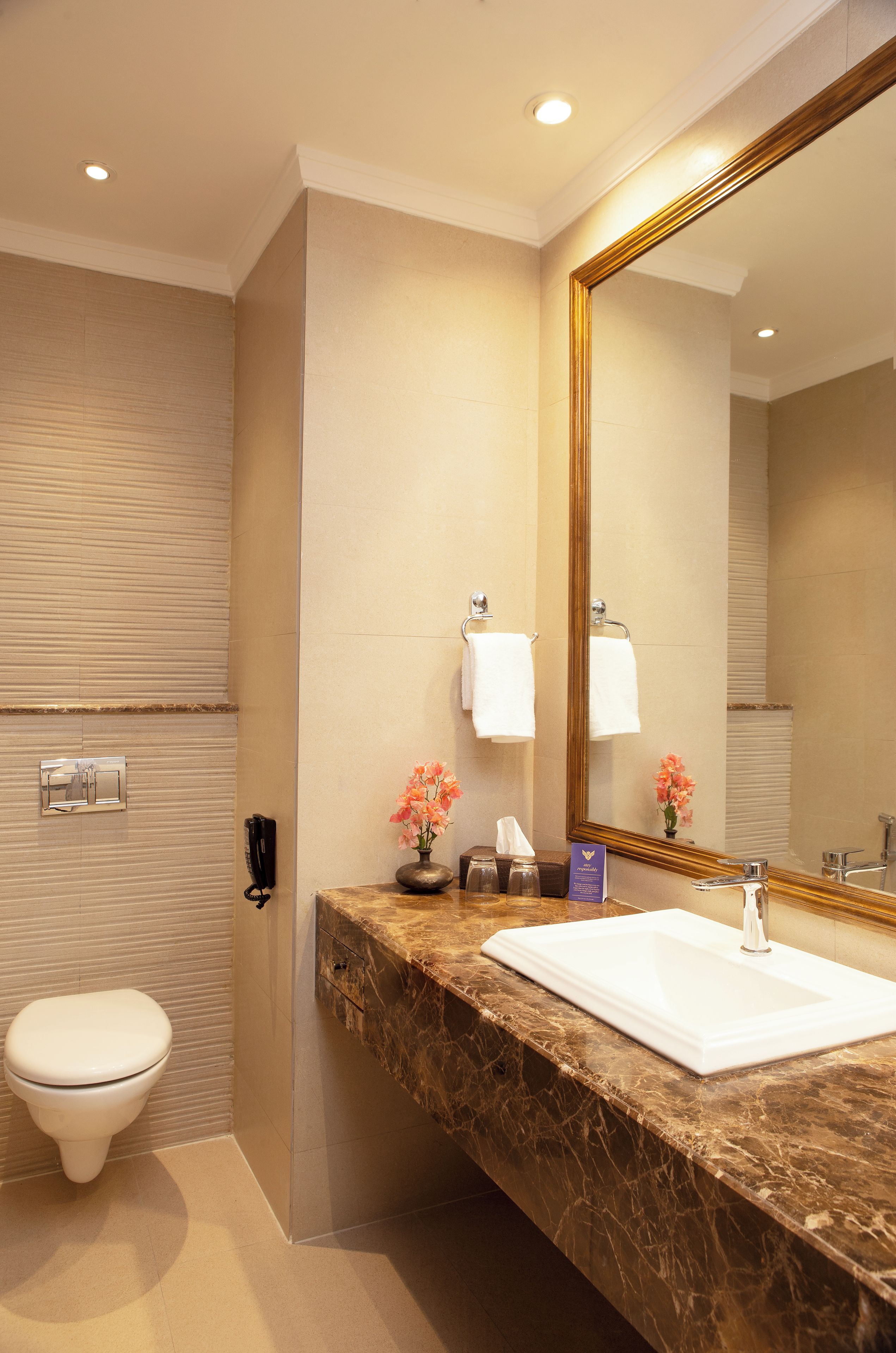 Un baño de hotel con tocador de mármol, lavamanos blanco, espejo grande y retrete.