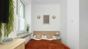 Une chambre d’hôtel comprenant un lit, une table de chevet, une horloge numérique, une fenêtre avec stores, une œuvre d’art accrochée au mur et une plante en pot.
