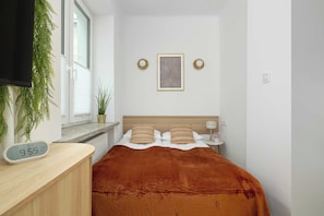 Een hotelkamer met een bed, nachtkastje, digitale klok, raam met jaloezieën, wanddecoratie en een bloempot met plant.
