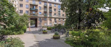Un edificio de varios pisos con una entrada central, rodeado de vegetación y un paseo empedrado.