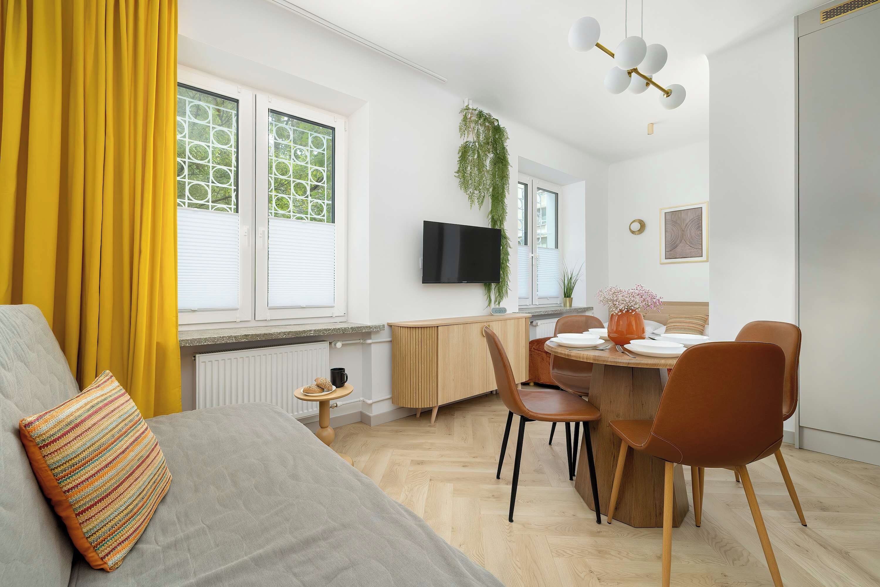 A modern living room with a sofa, a dining table with chairs, a flat-screen TV, and a large window with blinds.