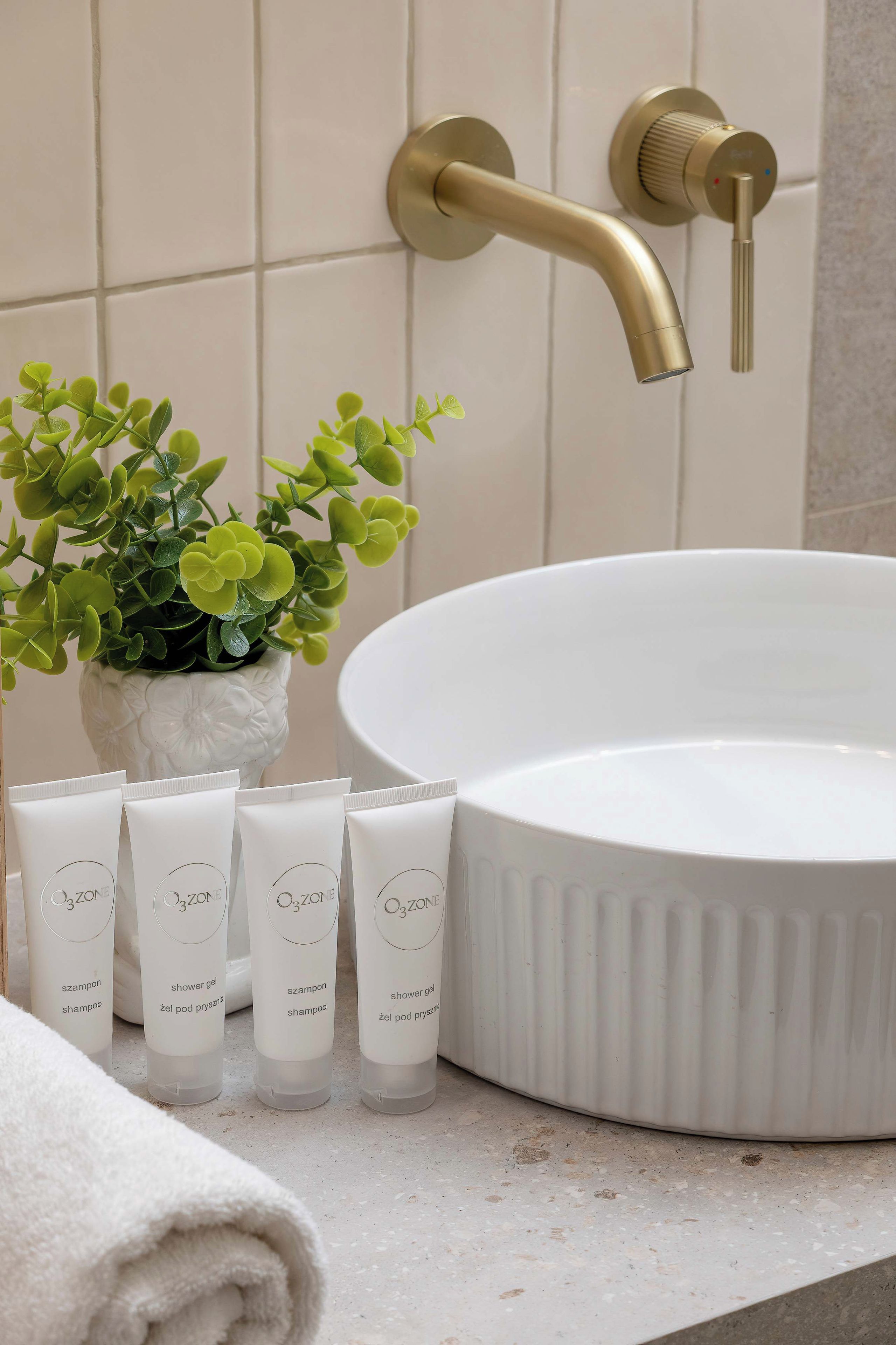 A bathroom with a white sink, gold faucet, and a potted plant. There are four bottles of O3ZONE shower gel and shampoo.