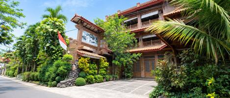 A traditional two-story building with a tiled roof, surrounded by lush greenery and a stone pathway leading to the entrance.