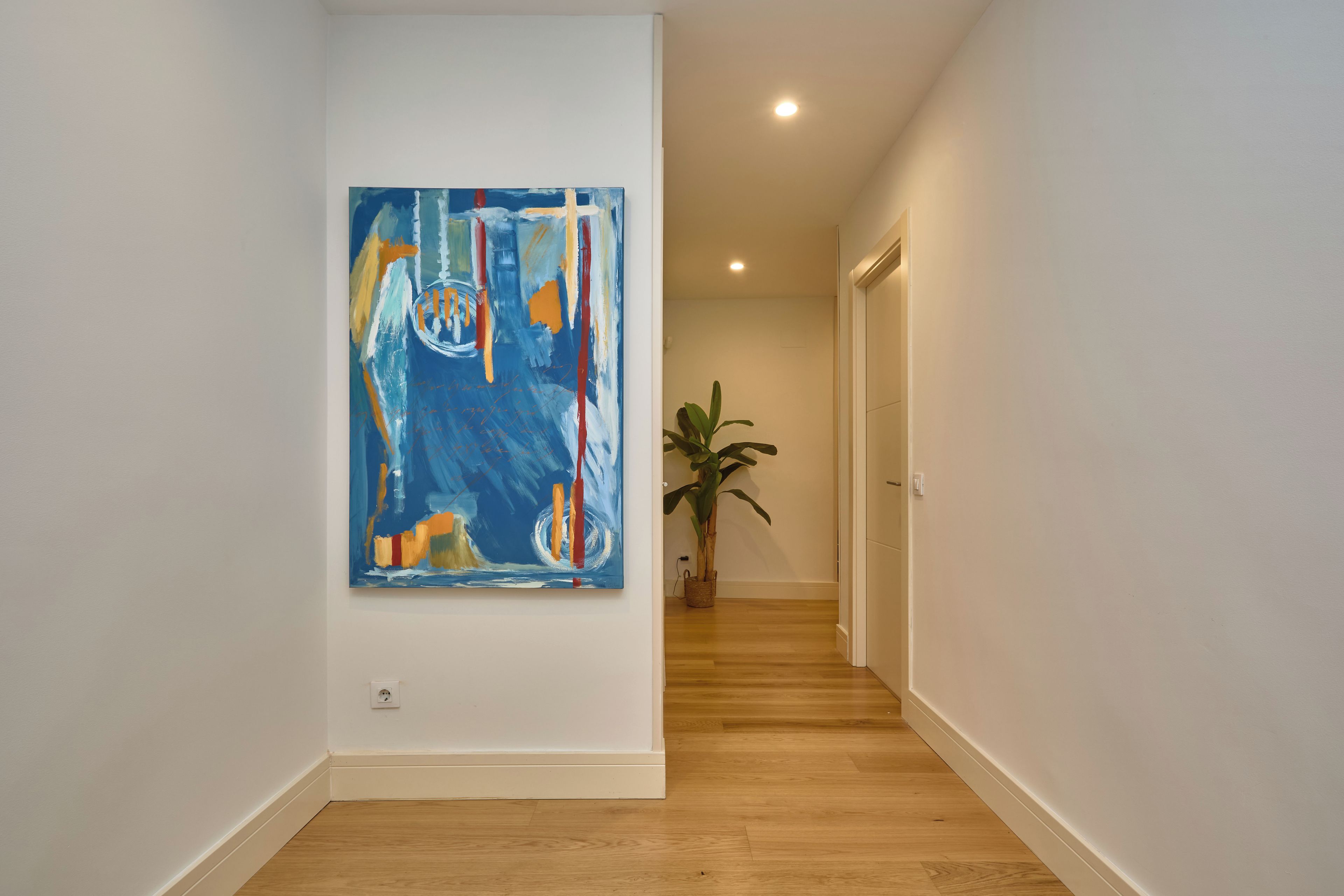 Ein Flur mit einem großen, abstrakten Gemälde an der linken Wand, einer Zimmerpflanze rechts und Holzböden.