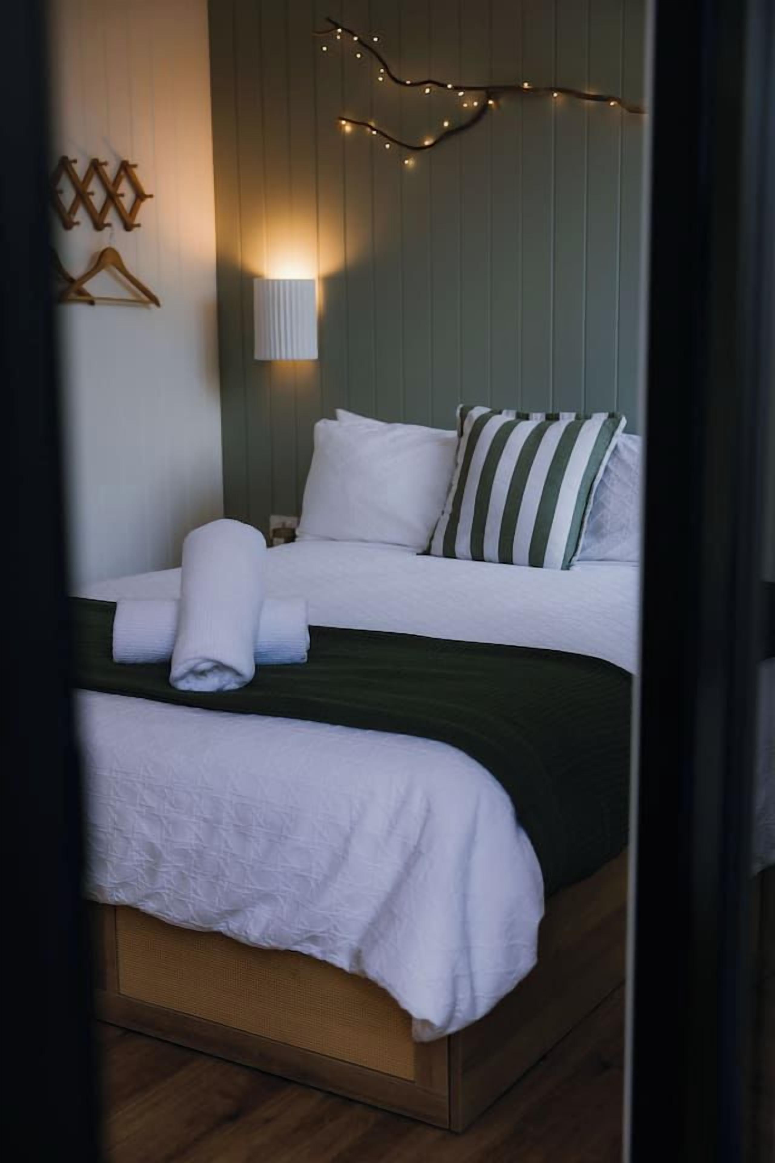 A double bed with white linens and a green bedspread, a bedside lamp, and string lights on the wall.