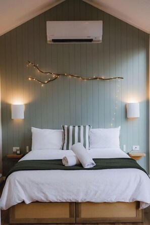 A bedroom with a bed, two bedside tables, a wall-mounted air conditioner, and string lights.
