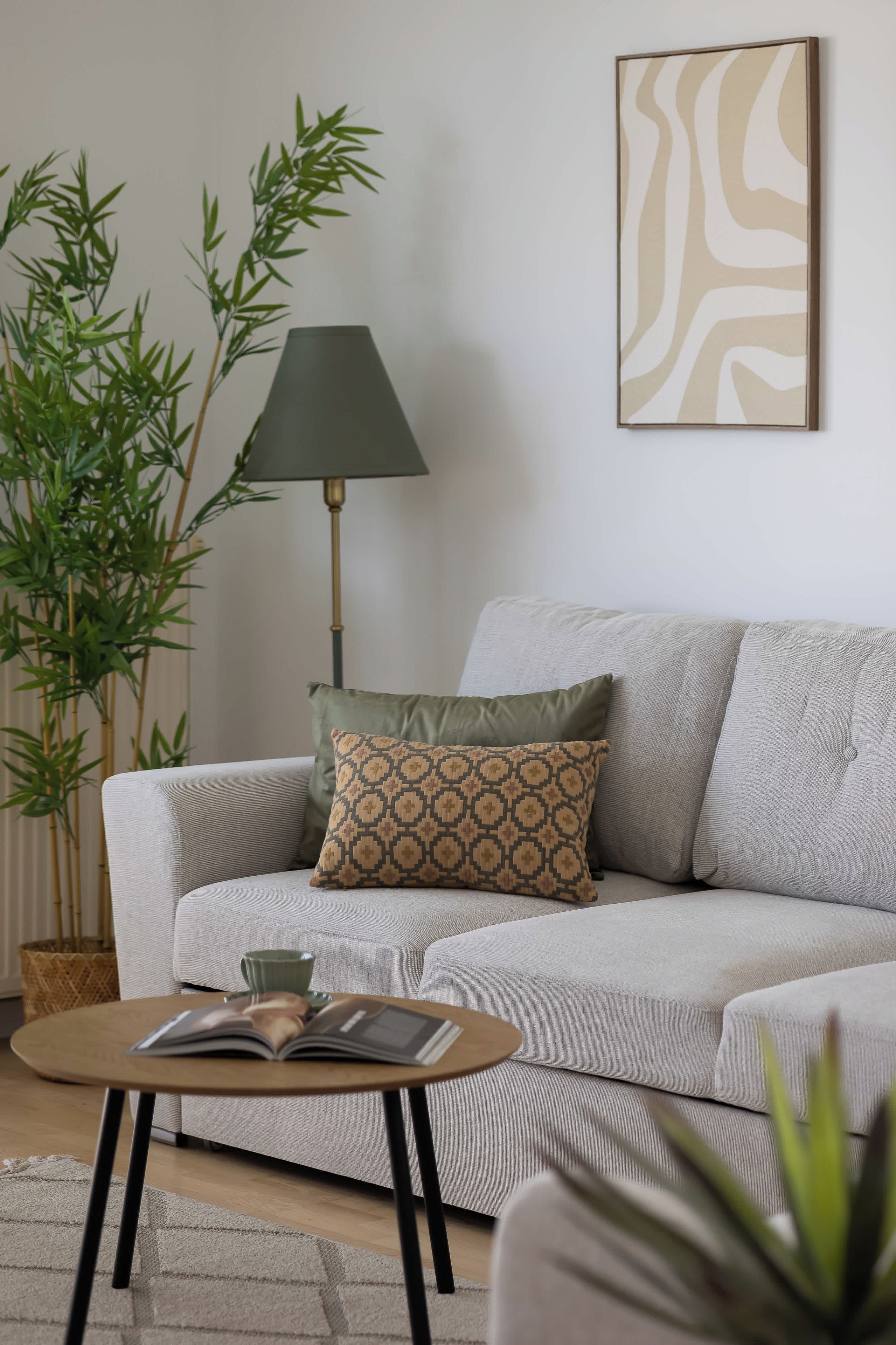 A living room with a grey sofa, a patterned throw pillow, a floor lamp, a green plant, and a framed abstract artwork on the wall.