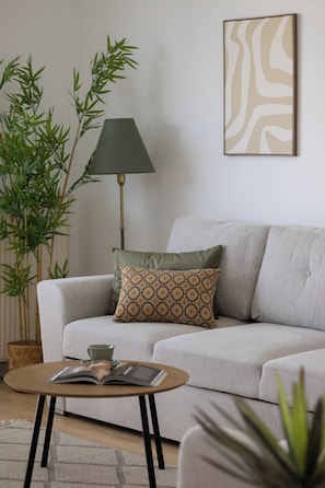 A living room with a grey sofa, a patterned throw pillow, a floor lamp, a green plant, and a framed abstract artwork on the wall.