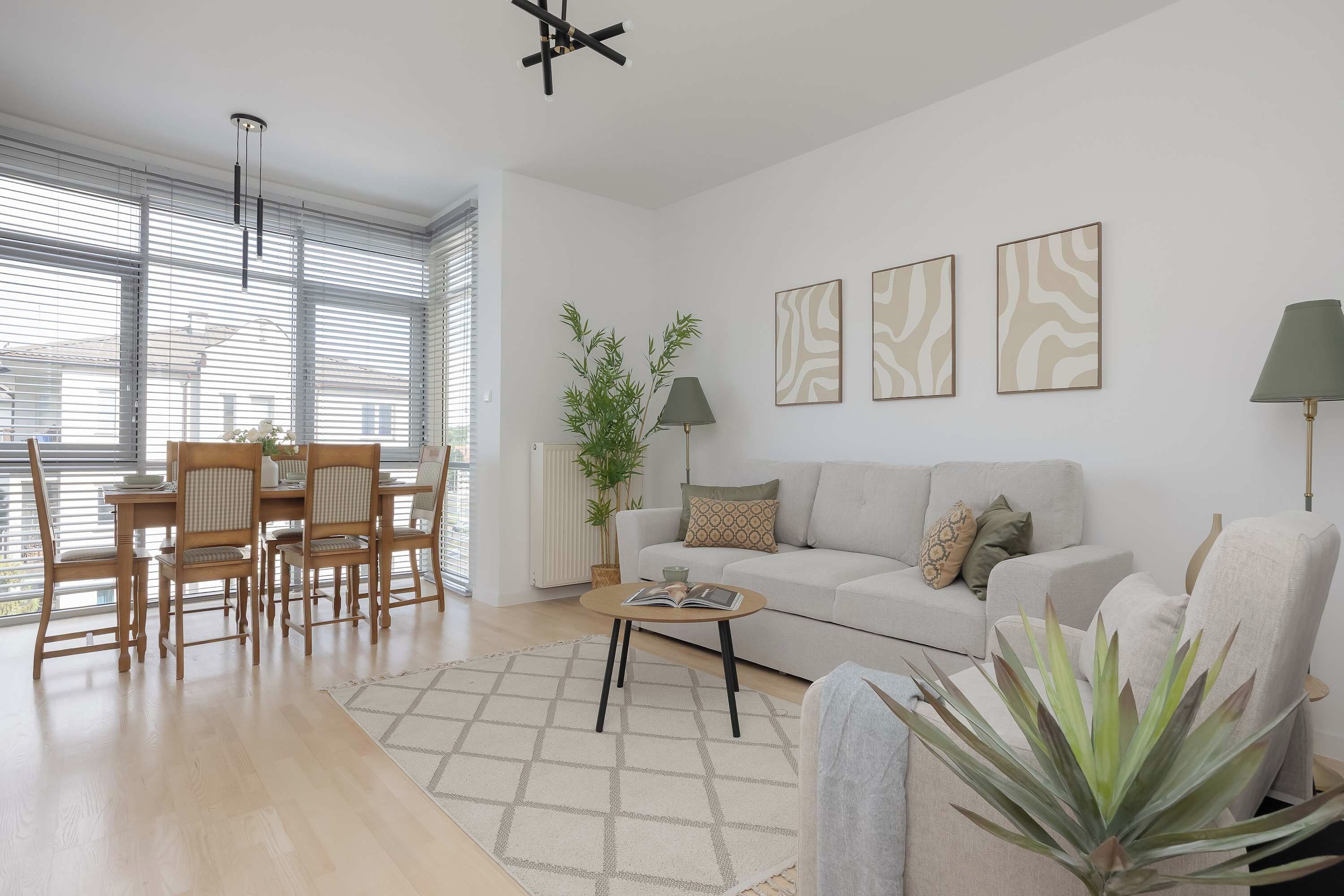 A modern living room with a sofa, a dining table with chairs, and a large window with blinds.