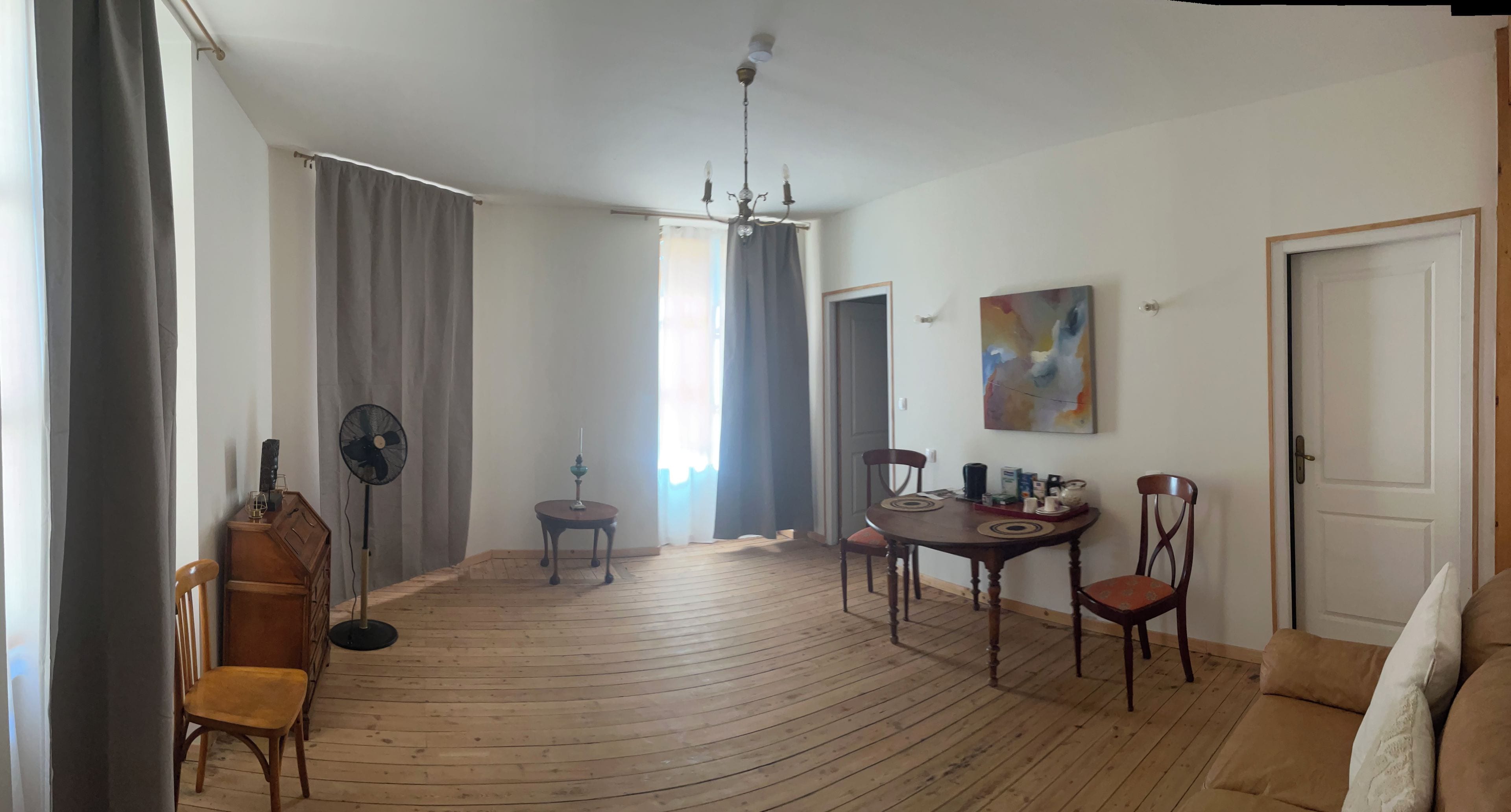 Een kamer met een houten vloer, een eettafel met stoelen, een ventilatorkachel, een plafondlamp en een deur.