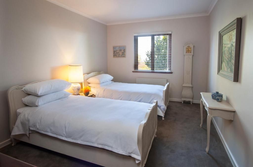 A bedroom with two beds, a bedside table with a lamp, a window with blinds, a radiator, and a clock on the wall.
