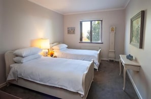 A bedroom with two beds, a bedside table with a lamp, a window with blinds, a radiator, and a clock on the wall.