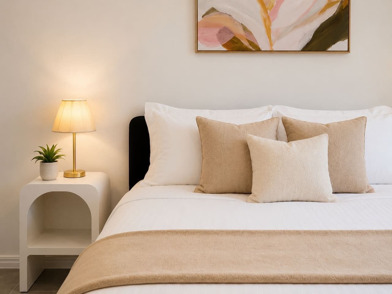 Ein ordentlich bezogenes Bett mit beiger Decke und Kissen, einer Nachttischlampe und einem kleinen Topfpflanzchen auf einem weißen Nachttisch.