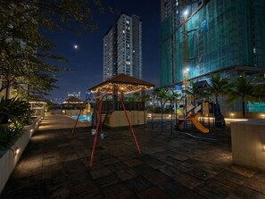 Un espacio exterior nocturno con un columpio, un pequeño quiosco y un tobogán, rodeado de edificios y palmeras.