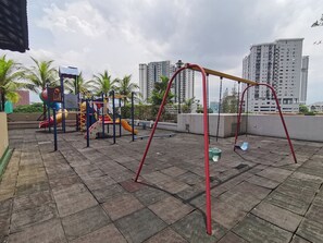 Un parque infantil con columpios y toboganes, rodeado de palmeras y rascacielos al fondo.