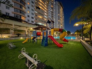 Zona de juegos al aire libre con toboganes y equipo de ejercicio, rodeada por una piscina y palmeras.