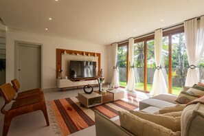 Una sala de estar moderna con un televisor de pantalla plana, muebles de madera, un tapete estampado y ventanas grandes con cortinas.