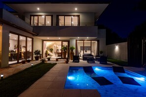 Una casa moderna de dos pisos con piscina, mobiliario exterior y un interior bien iluminado.