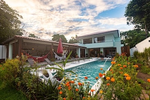 A pool area with a modern house, outdoor seating, and a hammock.